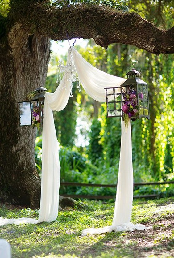 Rustic gets whimsical with this wedding altar consisting of draped fabric, lanterns filled with rich blooms, and a crystal chandelier