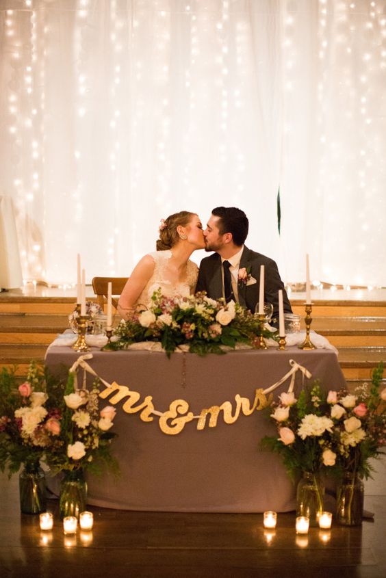 Twinkle light backdrop of kissing bride and groom at sweetheart table with gold mr and mrs banner and candles gold candlesticks